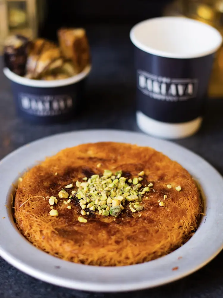 Round kunefe on a metal plate topped with crushed pistachios, next to a branded coffee cup