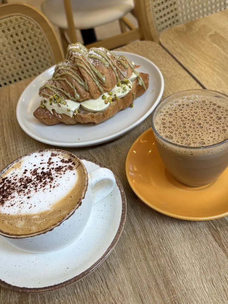 Pistachio cream croissant on a white plate beside a cappuccino and a glass of karak chai